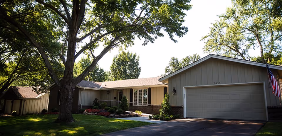 Ranch-style single-story home with attached two-car garage, driveway, front lawn and large shade trees.