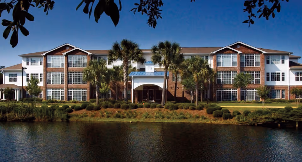 Front exterior view of a three-story senior living facility building named The Lakes at Litchfield, featuring large windows, palm trees, and landscaping in front of a pond under a clear blue sky.