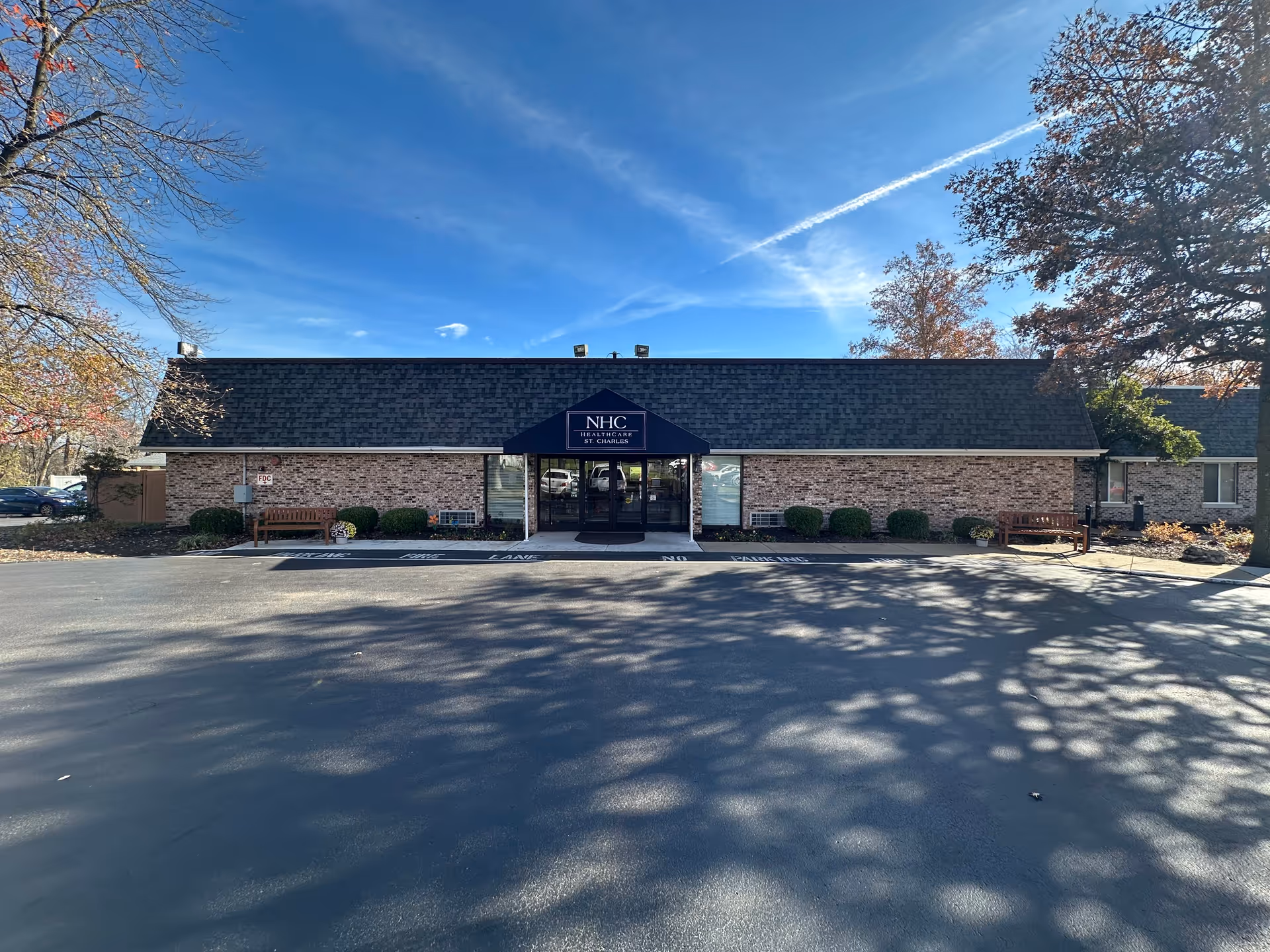 Front view of a single-story brick senior healthcare building with a blue awning entrance and a paved parking lot under a clear blue sky.