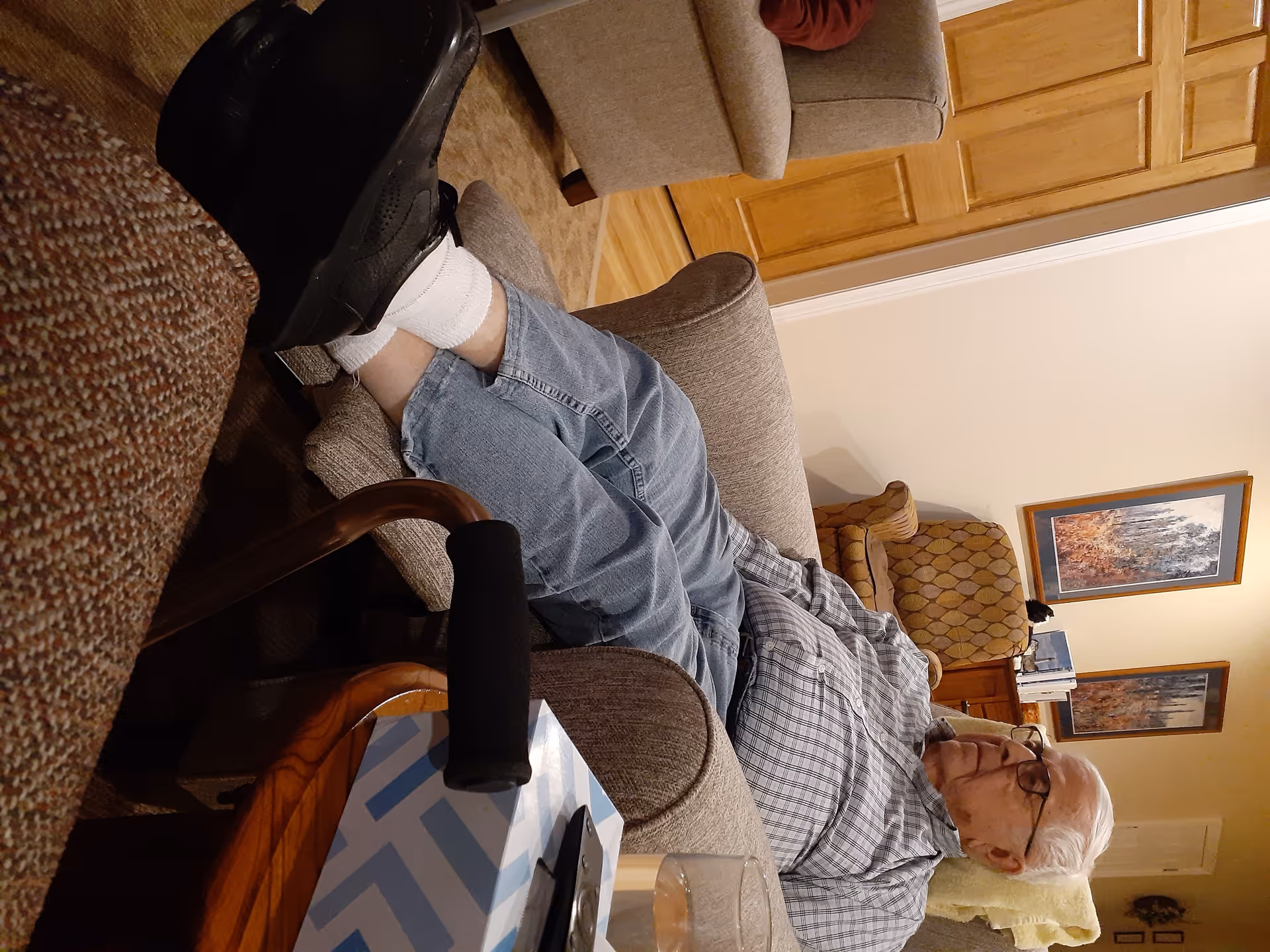 An elderly man with white hair and glasses is reclining on a beige armchair in a cozy living room. He is wearing a checkered shirt, blue jeans, white socks, and black shoes. Nearby is a wooden side table with a tissue box and a glass of water. The room has warm lighting, wooden furniture, and framed artwork on the walls.