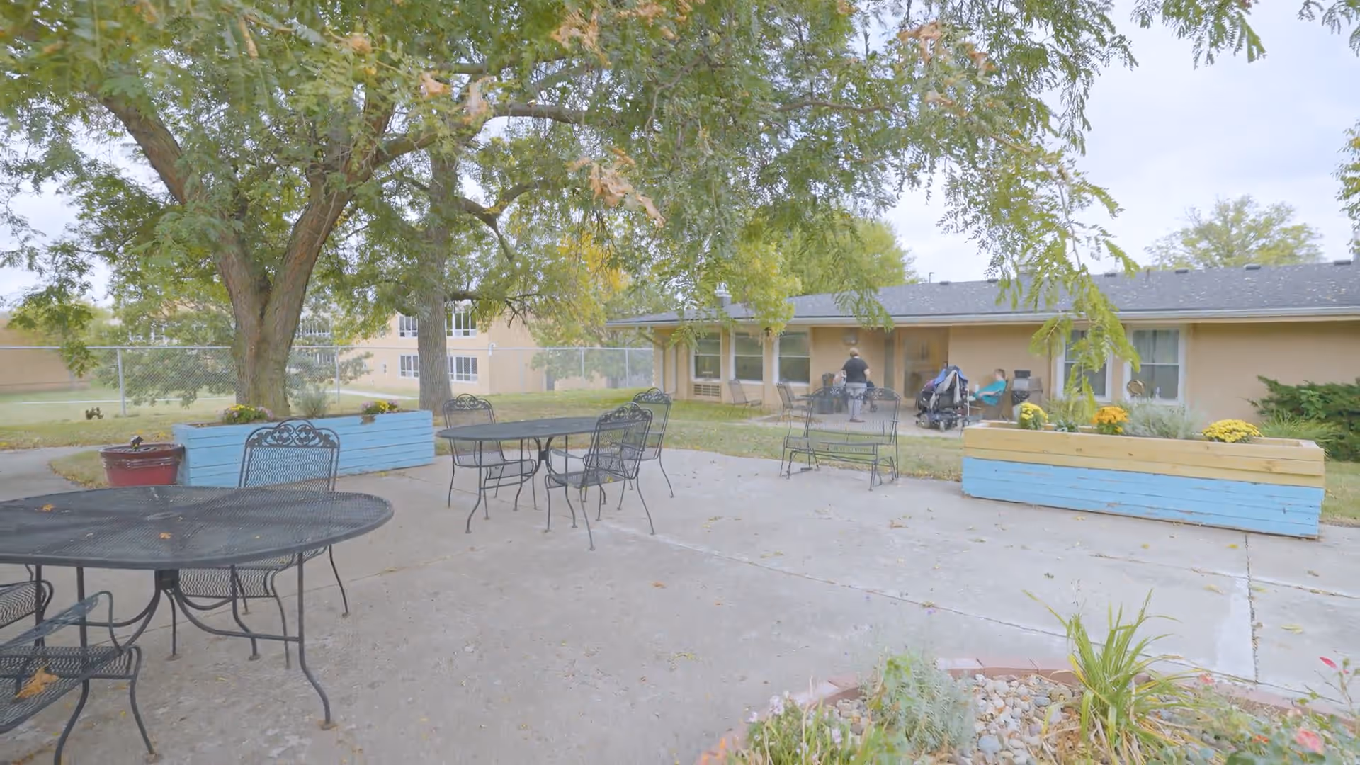 Patio outside a one-story senior living building with metal tables and chairs, planters and trees, and a few people near the entrance.