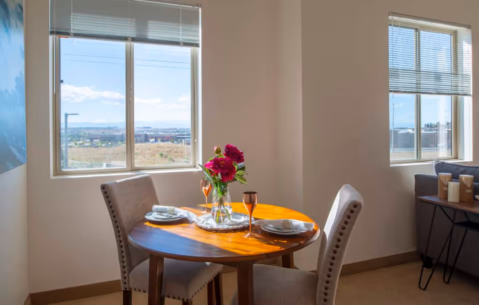 A small round wooden dining table set for two with plates, napkins, and copper-colored glasses. A vase with red flowers is in the center of the table. The table is positioned near two large windows with blinds, letting in natural light and showing an outdoor view. Part of a gray sofa and a side table with candles are visible in the background.