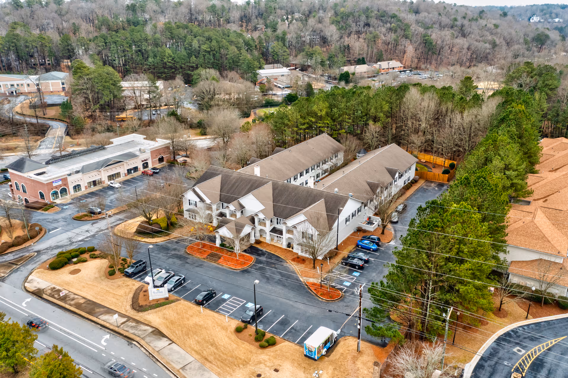 Aerial view of TerraBella Roswell senior living facility showing multiple connected buildings surrounded by trees and parking lots with several cars parked. The area includes landscaped grounds and nearby commercial buildings.