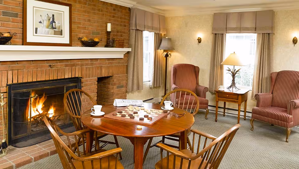 Cozy living room with a lit brick fireplace, a round wooden table with a checkers game and two cups, surrounded by four wooden chairs. Two striped armchairs flank a small wooden side table with a lamp, and two windows with beige curtains allow natural light into the room.