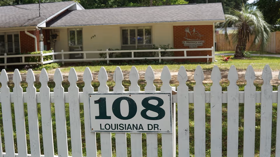 White picket fence with an address sign reading "108 Louisiana Dr." in front of a one-story house displaying a "FL Memory Lane" sign.