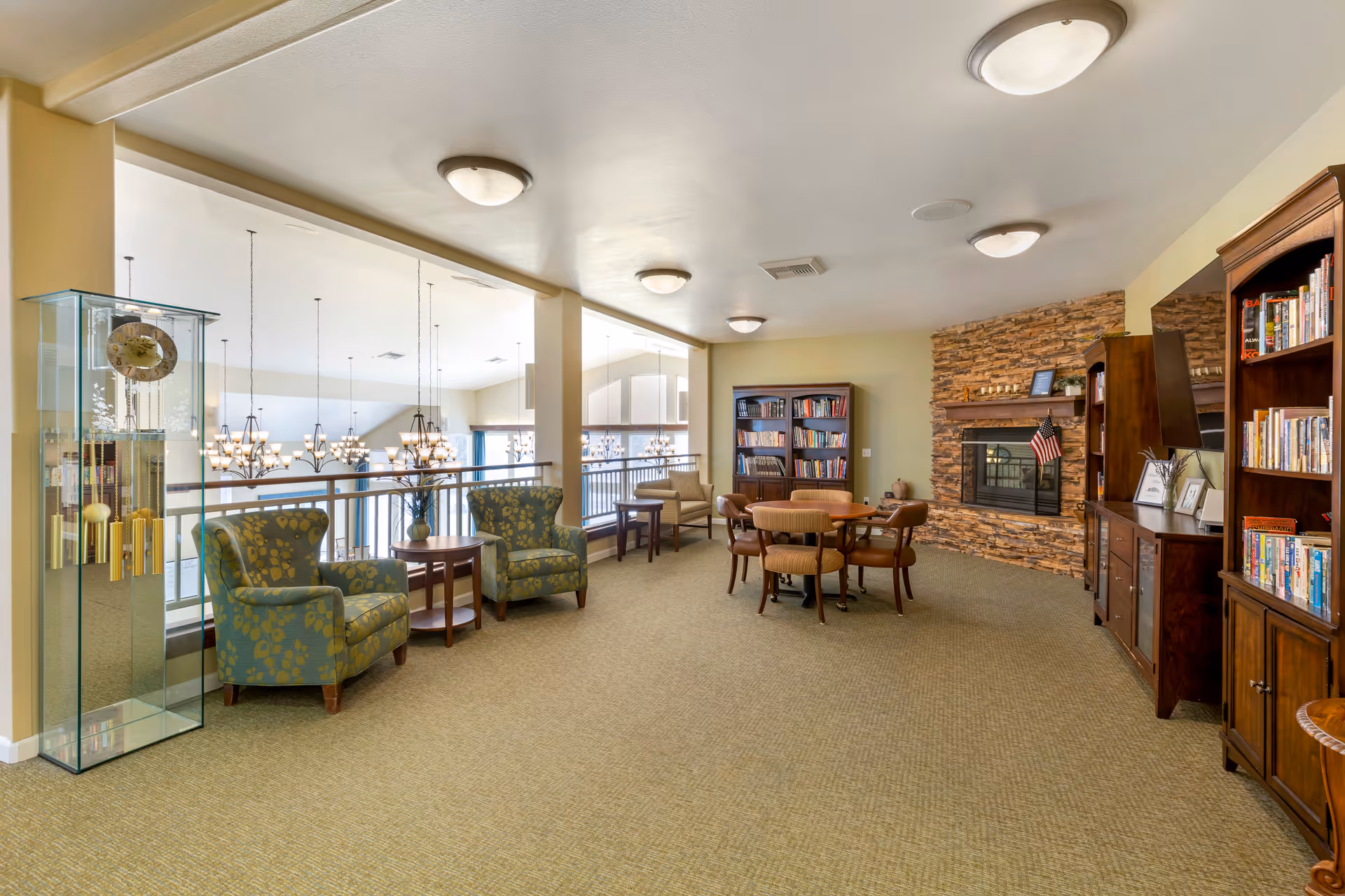 A spacious senior living facility common area with beige carpet and light yellow walls. The room features two patterned armchairs with a small round wooden table between them, a round table with four chairs, multiple wooden bookshelves filled with books, and a stone fireplace with an American flag on the mantel. Large windows overlook a lower level with hanging chandeliers.