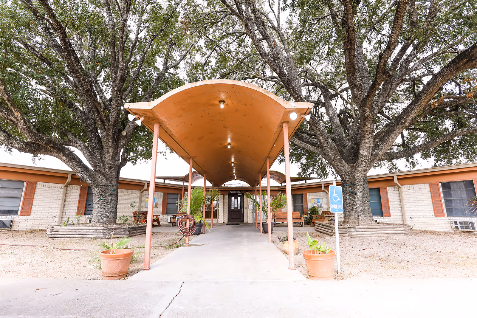 Entrance of Gulf Shores Rehabilitation and Healthcare Center with a covered walkway supported by metal poles, flanked by two large trees and potted plants. The building is a single-story structure with light-colored brick walls and brown window shutters. A handicapped parking sign is visible near the walkway.