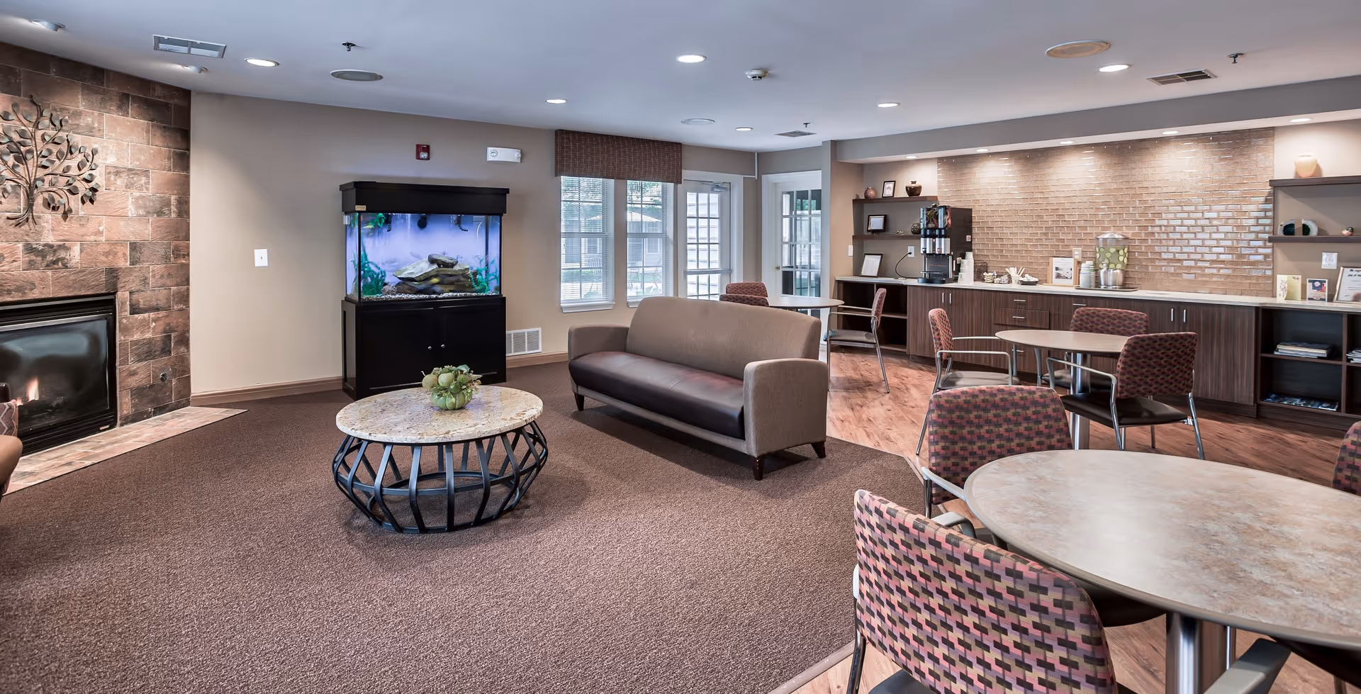 A cozy common area in a senior living facility featuring a brown carpeted floor, a stone fireplace with a metal tree wall decoration, a fish tank on a black stand, a brown sofa, round tables with patterned chairs, and a counter with a coffee machine and water dispenser under warm lighting.