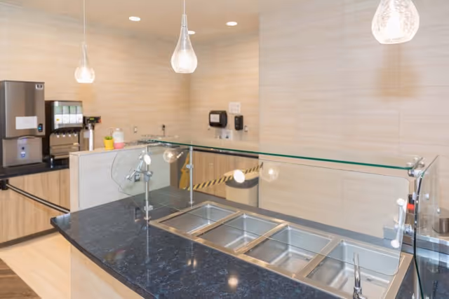 A clean and modern food service counter with a black countertop and multiple stainless steel food trays under a glass sneeze guard. In the background, there is a beverage dispenser and a wall with beige tiles, along with a soap dispenser and paper towel holder mounted on the wall.