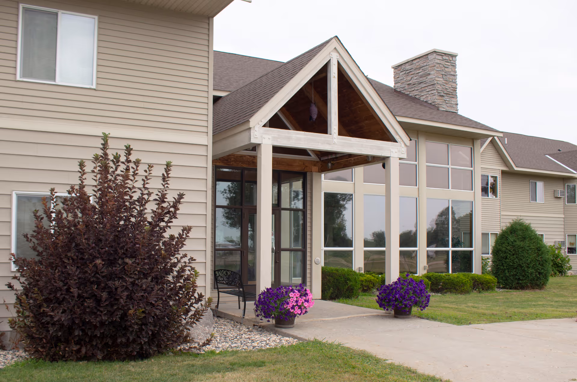 Exterior view of a senior living facility building with beige siding, large windows, and a covered entrance supported by white pillars. There are two flower pots with purple flowers near the entrance, a bench under the covered area, and landscaping including bushes and grass around the building.