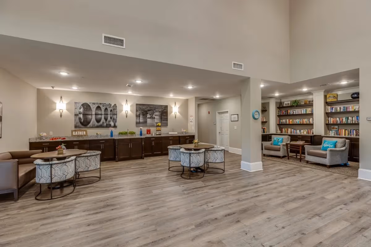Spacious senior living common area with seating, round tables, bookshelves and a long counter beneath wall art.