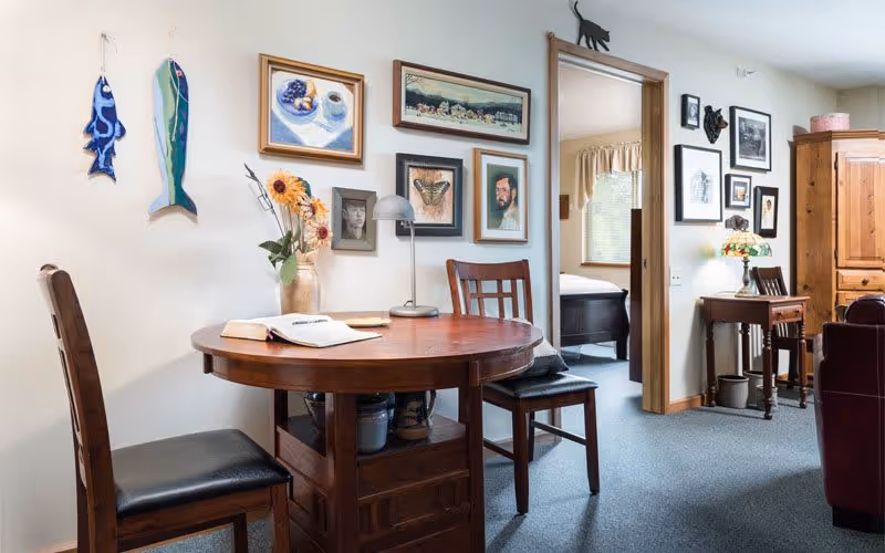 A cozy interior room with a round wooden table and two chairs. The table has an open book and a vase with sunflowers. The wall behind the table is decorated with various framed pictures and two hanging fish-shaped decorations. To the right, there is a doorway leading to a bedroom with a bed visible. Further right, there is a small wooden desk with a Tiffany-style lamp and more framed pictures on the wall. A wooden armoire and part of a leather chair are also visible.