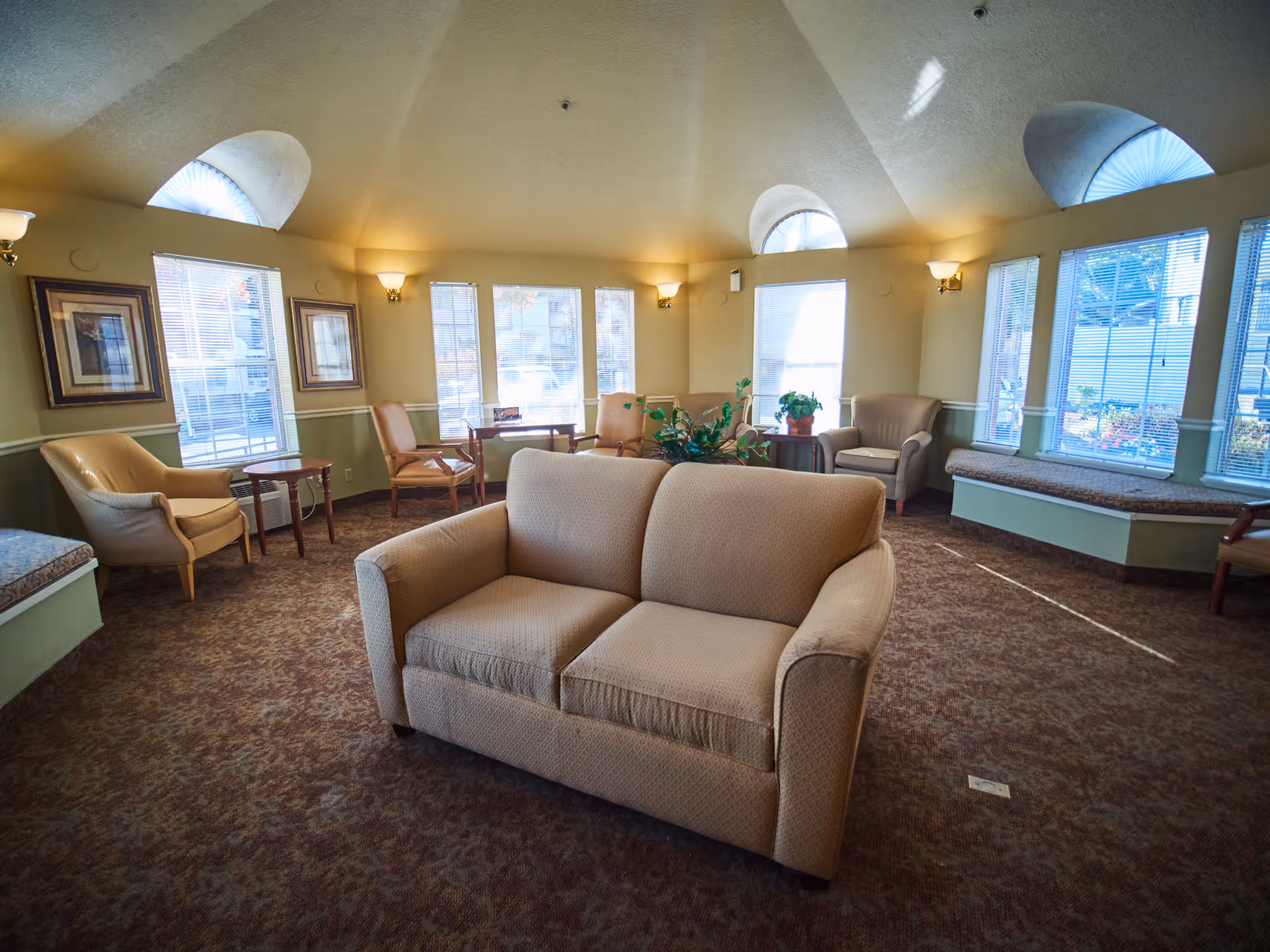 Bright communal seating room with a beige loveseat in the foreground, armchairs, window seats and arched windows.
