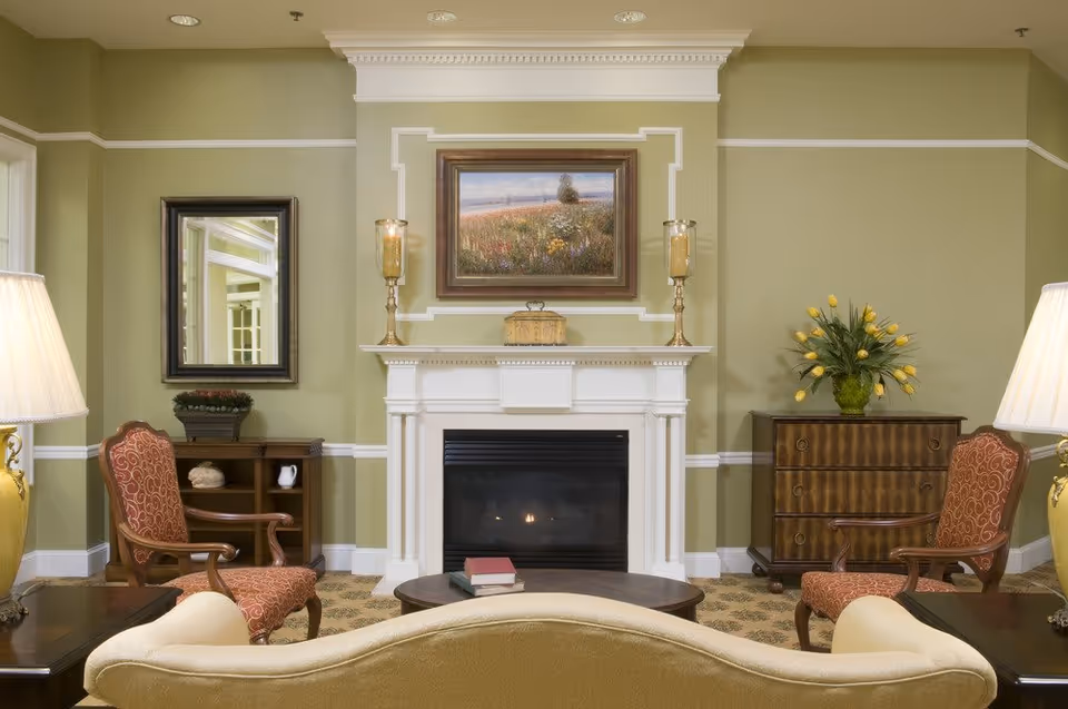 A cozy living room with a white fireplace centered on a green wall. Above the fireplace is a framed painting of a field with flowers. Two tall candle holders are placed on the mantel. On either side of the fireplace are wooden side tables with decorative items, including a vase with yellow tulips on the right. Two upholstered armchairs with red patterned fabric flank the fireplace. A beige sofa is in the foreground facing the fireplace, with a dark wooden coffee table holding two books in front of it. Two table lamps with white shades are positioned on side tables beside the sofa.
