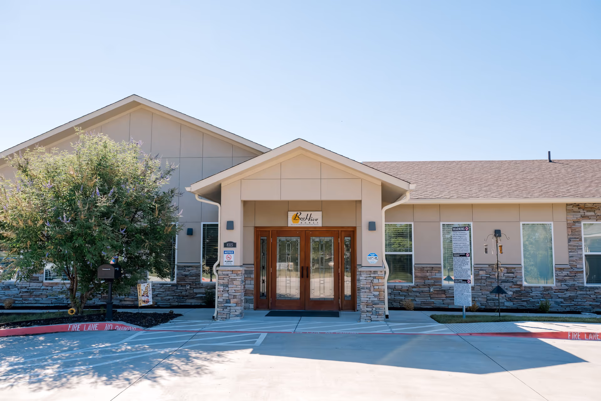 Front exterior view of BeeHive Homes of Rowlett building with a double wooden door entrance, stone and beige siding, a tree on the left side, and clear blue sky.