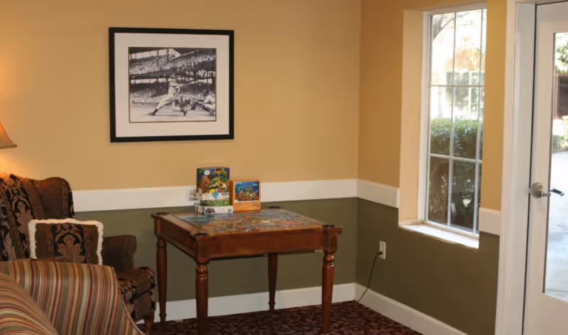 A cozy sitting area with a patterned armchair and a striped sofa next to a wooden table with puzzles on it. The walls are painted in two tones, beige on top and olive green on the bottom, with a white chair rail. A black and white framed photograph of a baseball game hangs on the wall. There is a window and a glass door letting in natural light.