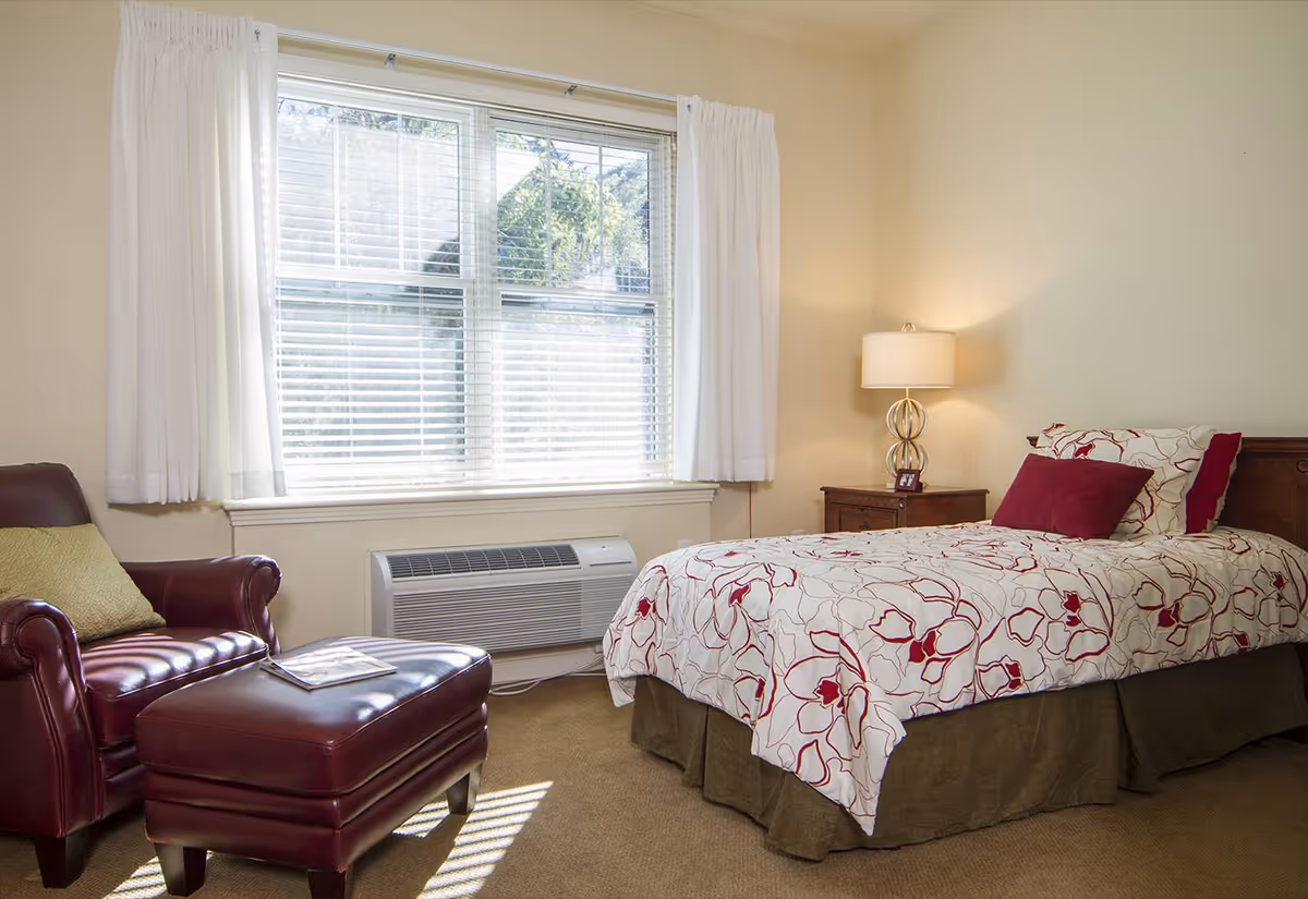 A cozy bedroom with a single bed covered in a floral patterned bedspread with red and white colors. Next to the bed is a wooden nightstand with a lamp and a small framed photo. There is a large window with white curtains letting in natural light. A dark red leather armchair with a matching ottoman and a green pillow is positioned near the window. The room has beige walls and carpeted floor.