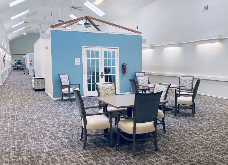 Large bright common room with several tables and chairs in front of a blue room with double French doors under a high skylit ceiling.