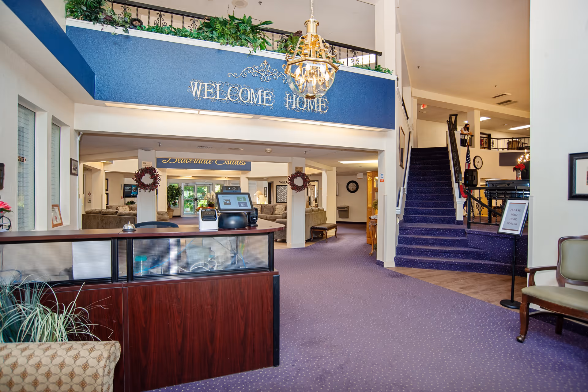 Interior view of a senior living facility lobby with a reception desk in the foreground, a blue carpeted floor, a staircase leading to an upper level, and a large sign that says 'WELCOME HOME' above the entrance to a common area with seating and plants.