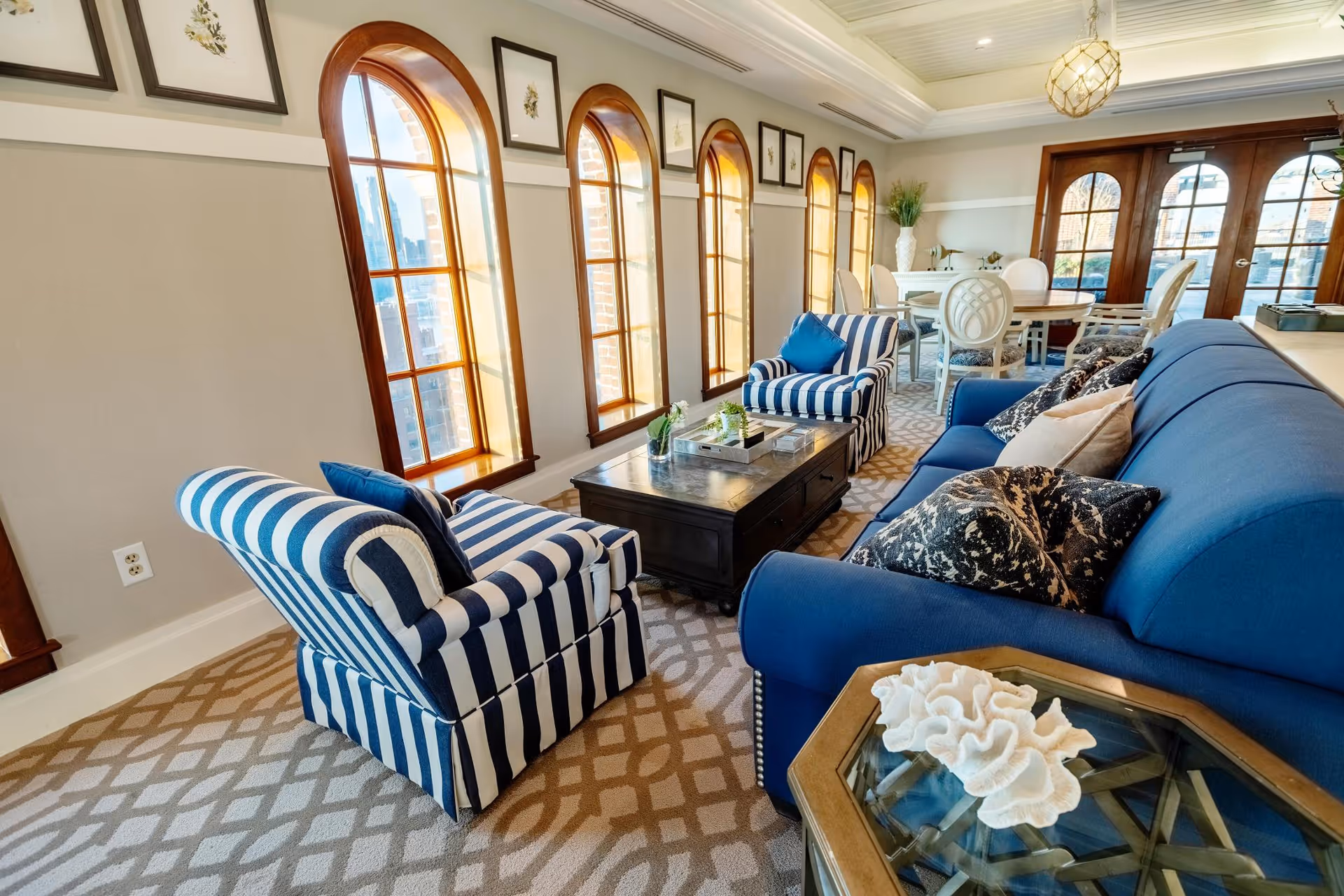 Sunlit common living room with a blue sofa, striped armchairs, coffee table and arched windows.