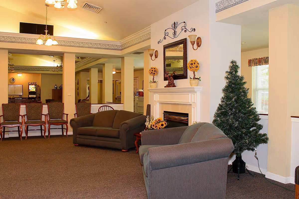 A cozy living room area in a senior living facility featuring two sofas, a fireplace with decorative items and a mirror above it, a small artificial Christmas tree, and several chairs lined up against the wall. The room has warm lighting and a carpeted floor.