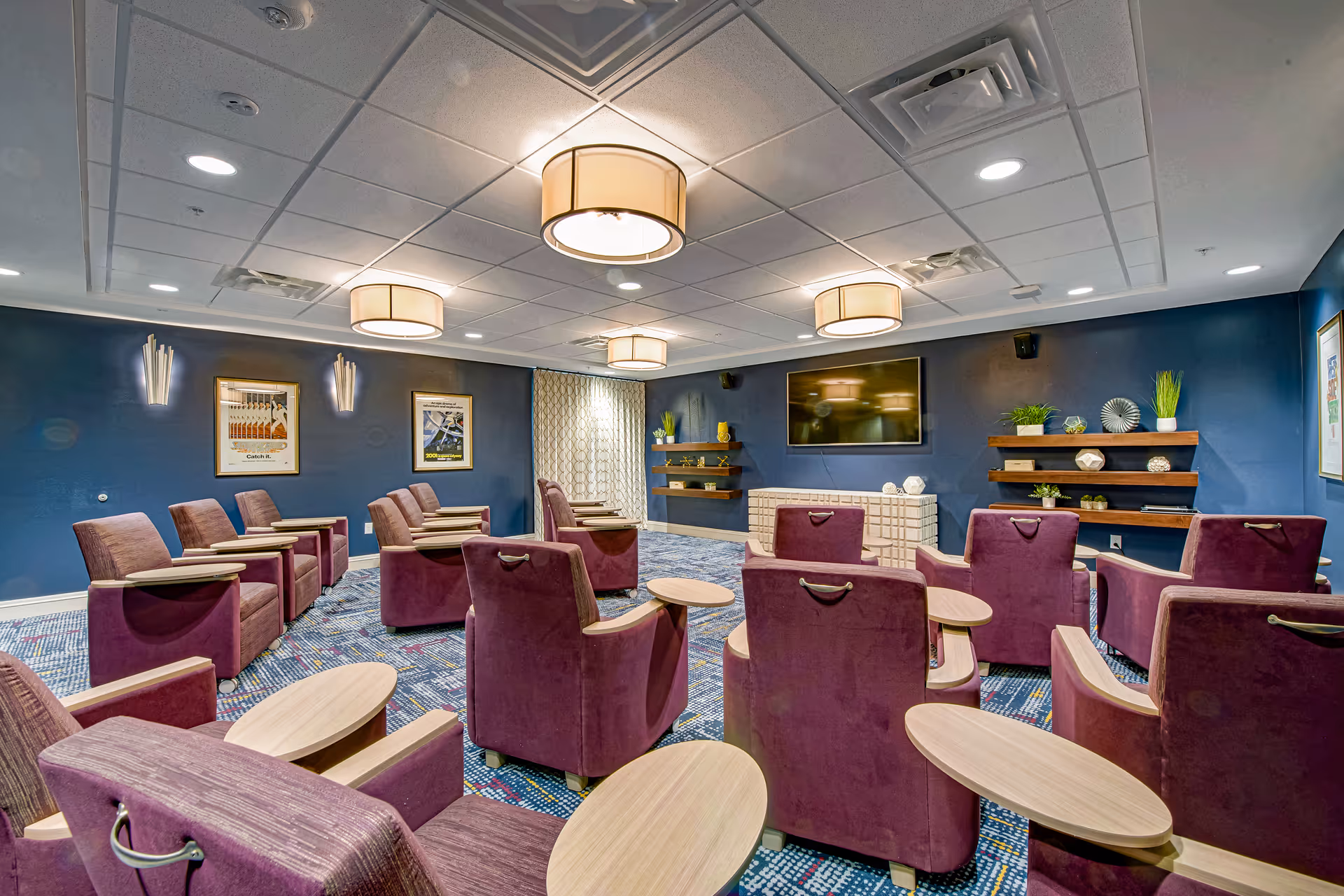 A cozy media room with multiple rows of purple armchairs equipped with small attached wooden tables, facing a large flat-screen TV mounted on a dark blue wall. The room has patterned carpet flooring, modern ceiling lights, and decorative shelves with plants and ornaments on the walls.