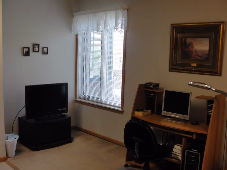 A small room with a window covered by white blinds and a lace valance. There is a black TV on a stand in the corner, a trash can beside it, and a wooden desk with a computer monitor, keyboard, and desk lamp. A black office chair is in front of the desk. The walls have a few framed pictures and a painting.