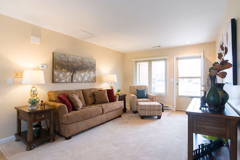 A bright living room with a brown sofa, patterned armchair, side tables with lamps, wall art, and a console table near a windowed door.