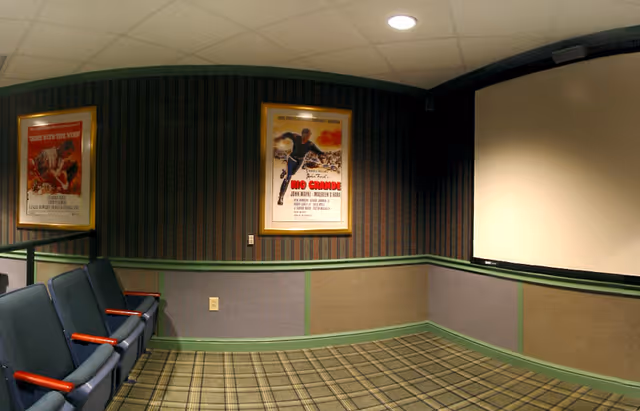 Small theater-style interior with upholstered row seating facing a projection screen, framed movie posters on paneled walls, and a patterned carpet.