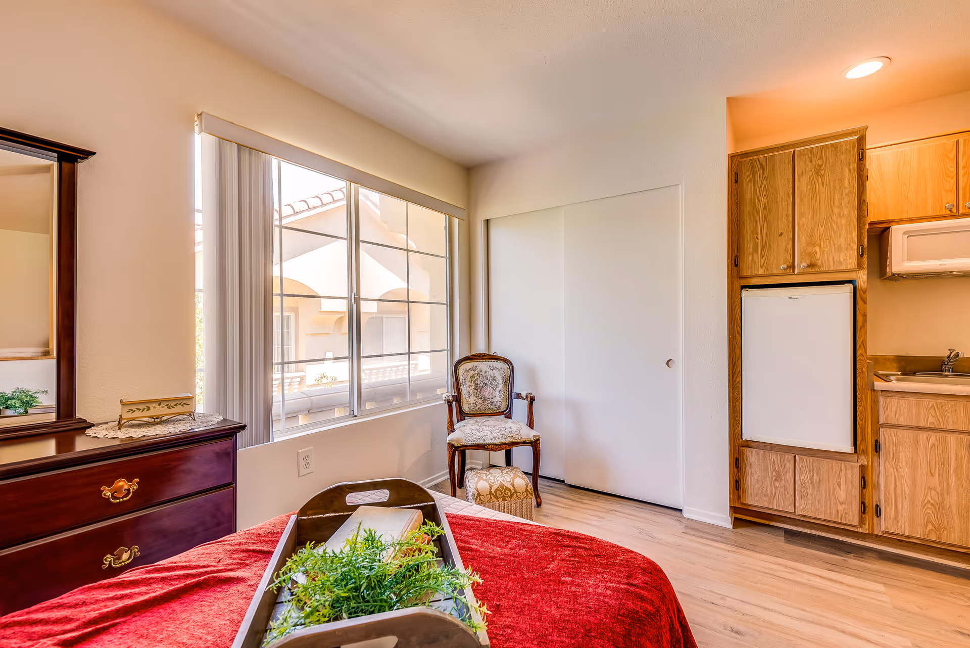 A bright bedroom with a large window showing an exterior view of a building. The room features a wooden dresser with a mirror, a bed with a red blanket and a wooden tray holding a plant and a book. There is an upholstered chair with a matching footstool near a white sliding closet door. To the right, there is a small kitchenette area with wooden cabinets, a mini refrigerator, and a microwave.