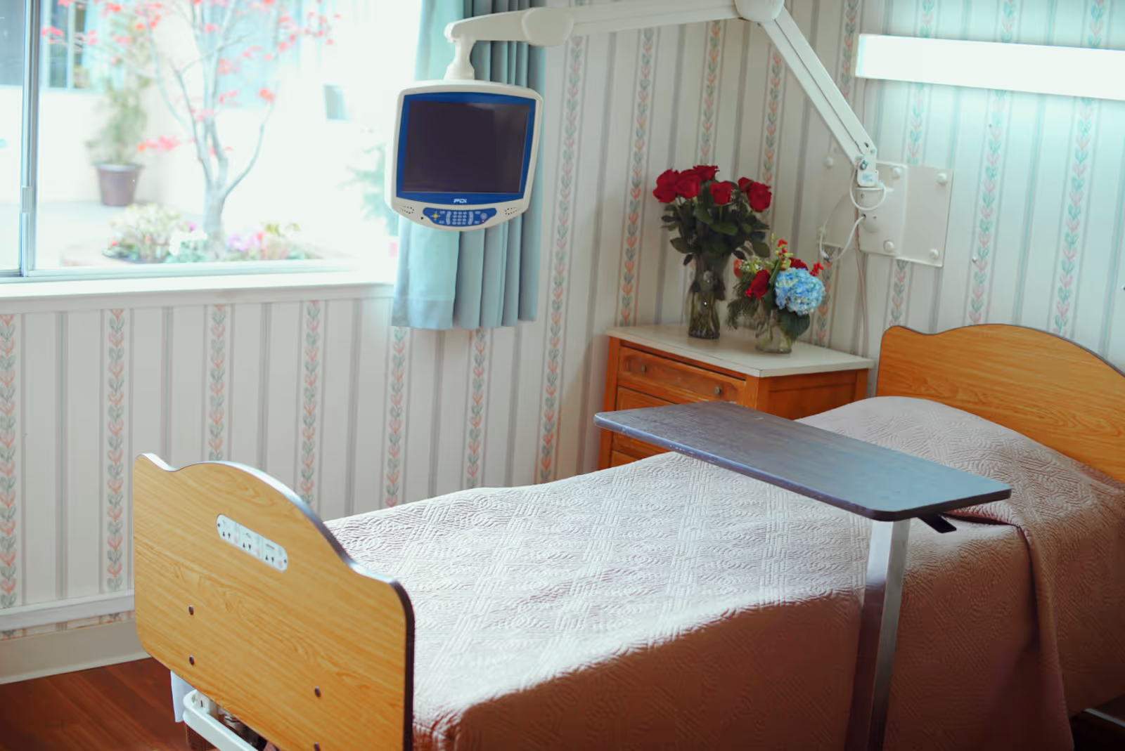 A hospital-style bed with a wooden headboard and footboard, covered with a beige quilted blanket. A movable overbed table is positioned over the bed. Next to the bed is a wooden nightstand with two vases of flowers, including red roses and blue hydrangeas. A medical monitor is mounted on an adjustable arm above the bed. The room has wallpaper with a vertical floral pattern and a window with light blue curtains letting in natural light.