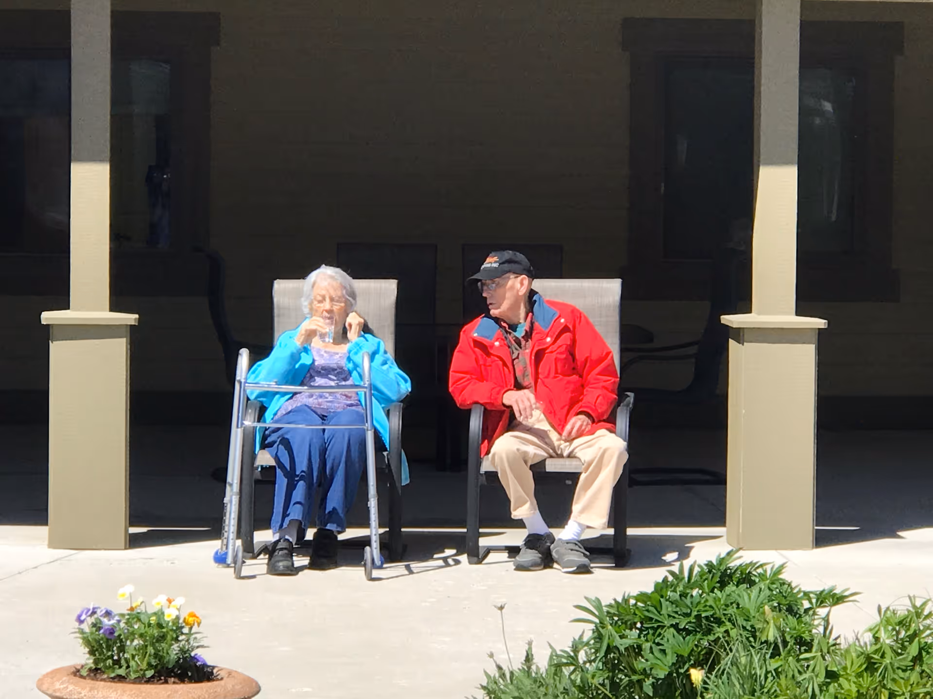 An elderly woman and an elderly man sitting outside on patio chairs under a covered porch. The woman is using a walker and drinking from a cup, while the man is wearing a red jacket and a black cap. There are flowers and greenery in the foreground.