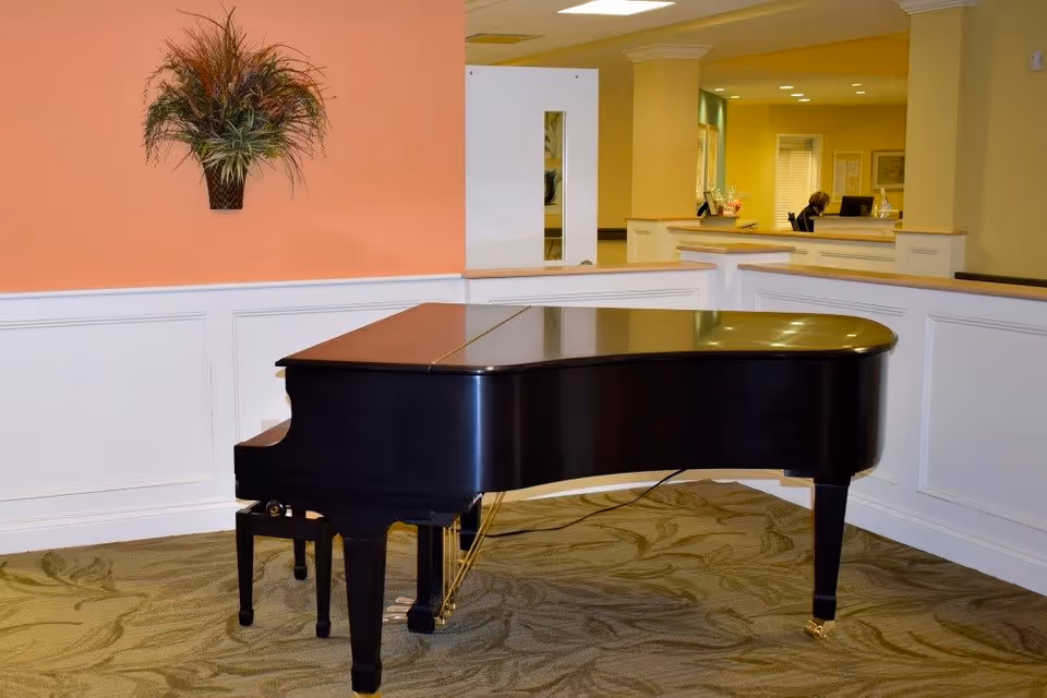 A black grand piano placed on a patterned carpet in a room with peach and yellow walls. There is a decorative plant mounted on the peach wall, and a reception desk area with a person working at a computer is visible in the background.