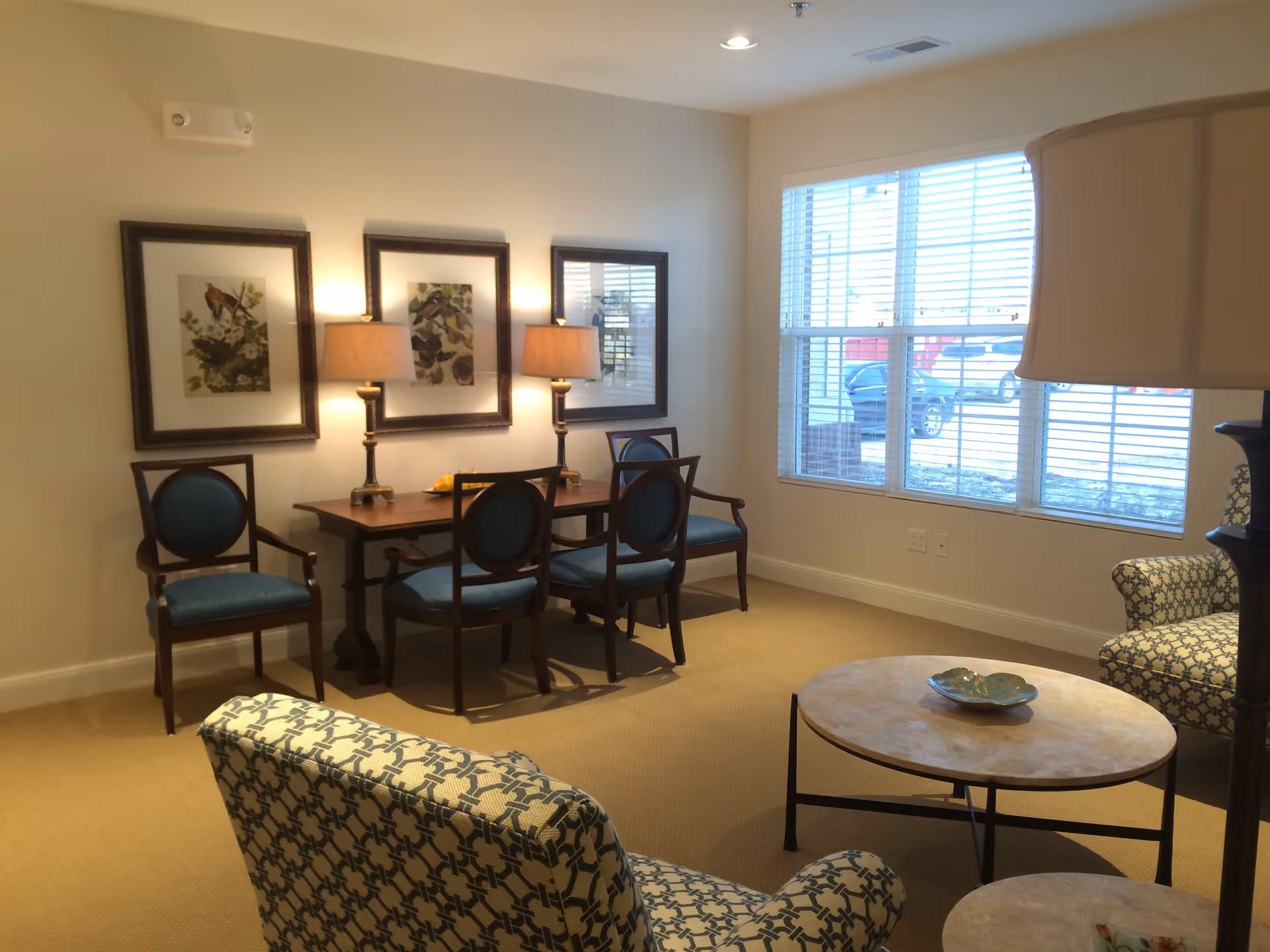 A cozy interior room with a small wooden table and four blue cushioned chairs against a wall with three framed pictures and two table lamps. There is a large window with blinds letting in natural light, and two patterned armchairs with a round coffee table in the foreground.