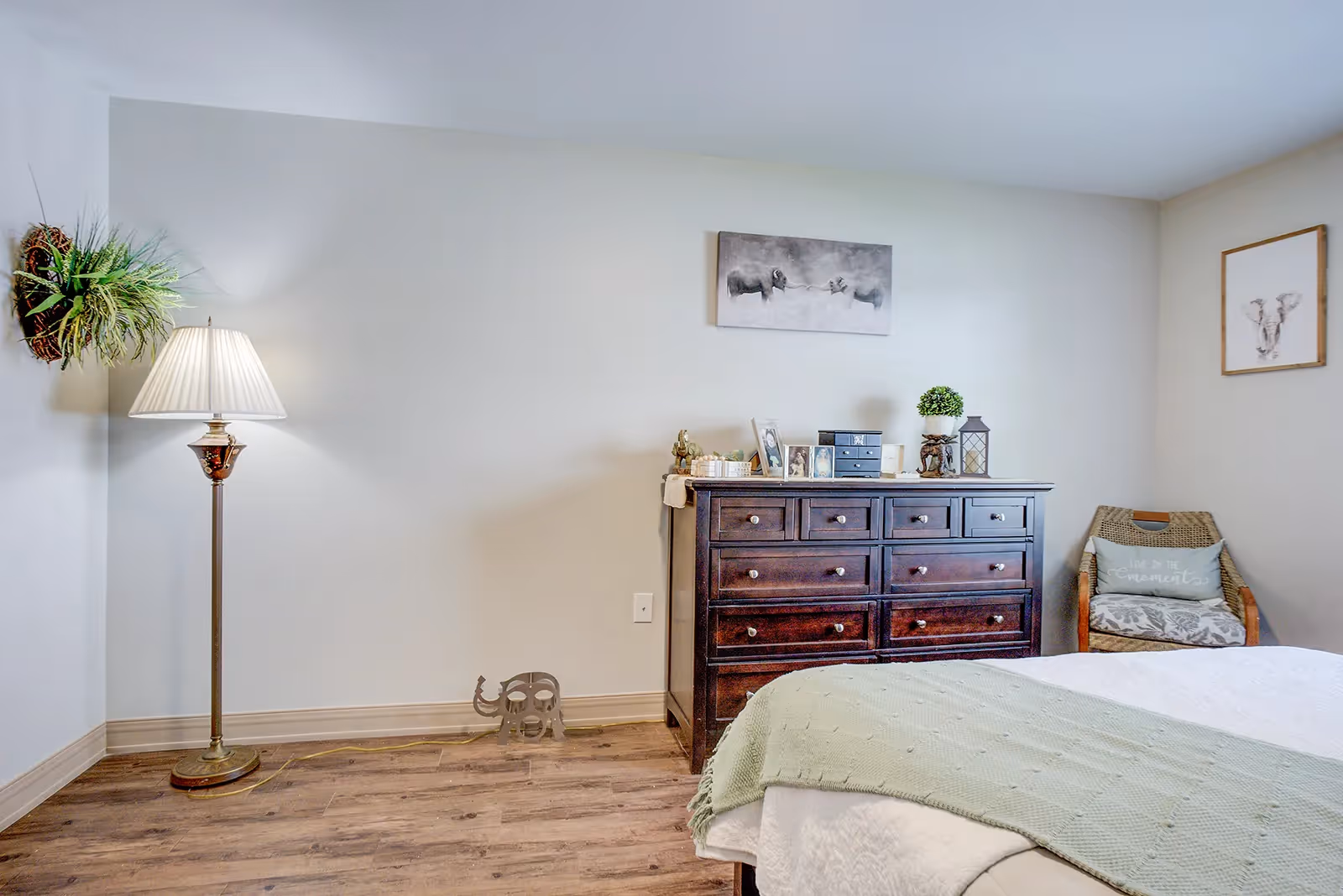 A cozy bedroom with a wooden dresser topped with framed photos, a small plant, and decorative items. A wicker chair with a cushion is placed in the corner next to the dresser. A floor lamp with a white shade stands near the wall, which is decorated with two pieces of animal-themed artwork. The bed has a light green blanket and white bedding, and the floor is wooden.