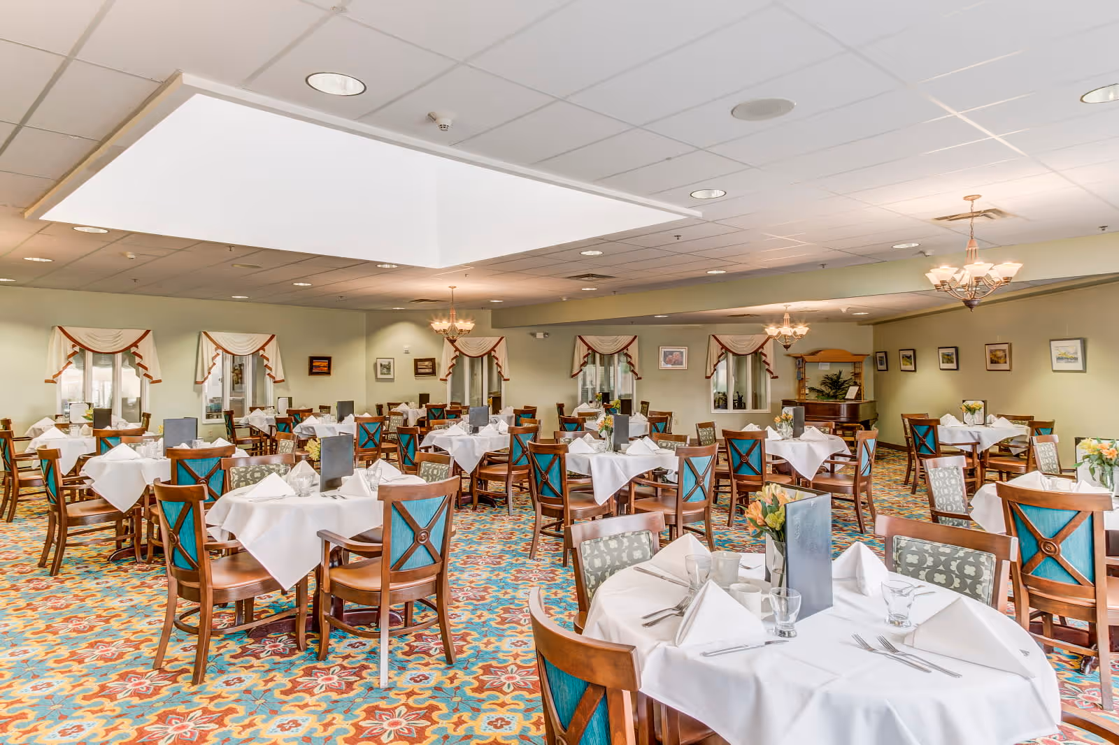A spacious dining room with multiple round tables covered with white tablecloths, each set with napkins, glasses, and silverware. The chairs have wooden frames with teal and patterned upholstery. The room features a colorful patterned carpet, soft green walls, framed artwork, and windows with valances. Ceiling lights and chandeliers provide warm lighting.