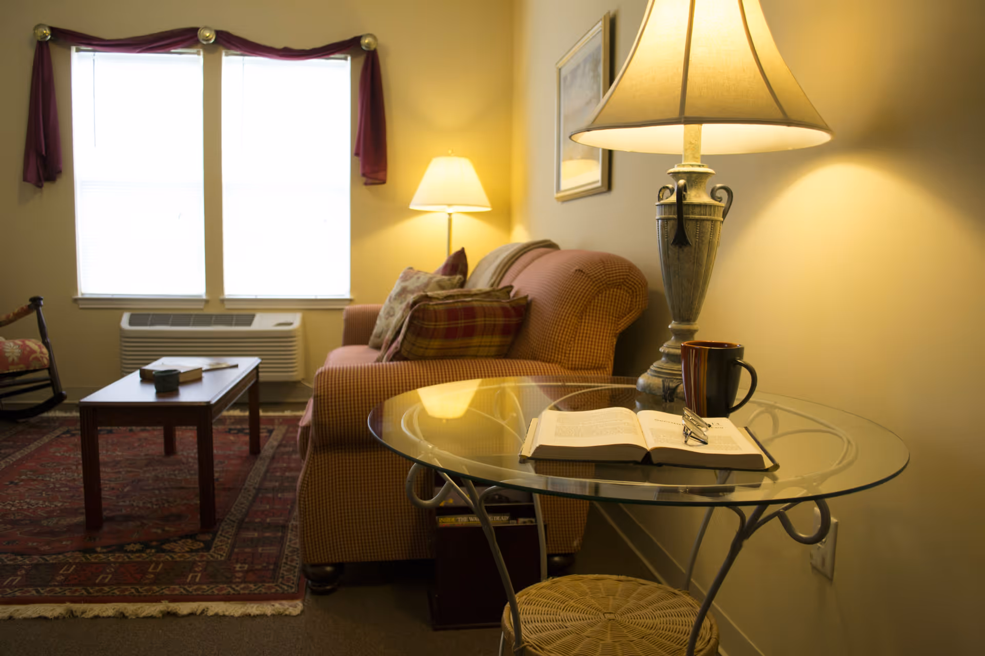 A cozy living room with a checkered sofa adorned with pillows, a glass side table with an open book, reading glasses, and a coffee mug. A lamp on the side table casts a warm light. In the background, there is a wooden coffee table, a patterned rug, a rocking chair, and two windows with purple curtains.