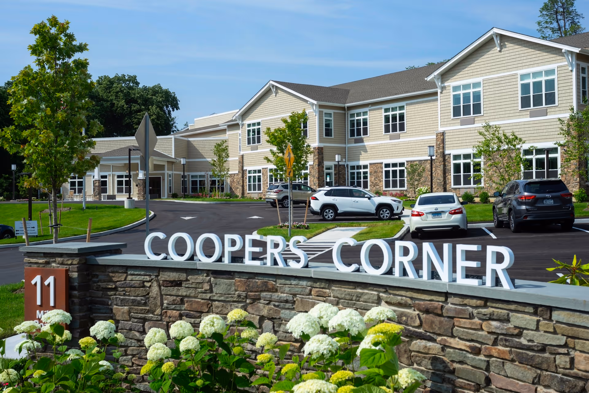 Exterior view of Monarch Coopers Corner facility showing a two-story building with beige siding and stone accents, a parking lot with several cars, green trees, and a stone sign with white letters spelling 'COOPERS CORNER'.
