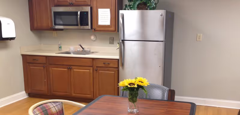 A small kitchen area with wooden cabinets, a stainless steel refrigerator, a microwave, and a sink. In the foreground, there is a wooden table with a vase of yellow sunflowers and chairs around it. The walls are light-colored and there is a paper towel dispenser mounted on the left wall.