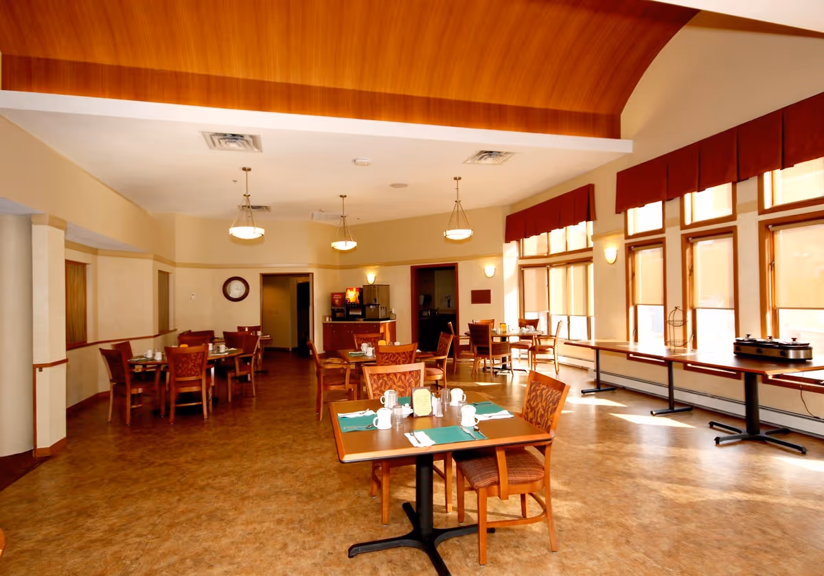 A well-lit dining room with several wooden tables and chairs arranged neatly. Each table is set with placemats, cups, and utensils. Large windows with red valances allow natural light to fill the room. The ceiling has a wooden arch design and hanging light fixtures. A beverage station is visible at the back of the room.
