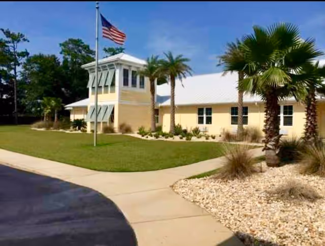 Exterior view of a single-story building with a light yellow facade and a white roof, surrounded by palm trees and landscaping with grass and rocks. An American flag is flying on a flagpole in front of the building under a clear blue sky.