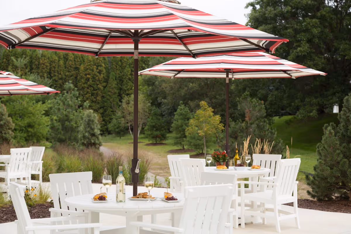 Outdoor patio area with white tables and chairs under red, white, and black striped umbrellas. Tables are set with wine glasses, bottles of wine, plates of food, and a small potted plant. The background features green trees and shrubs.