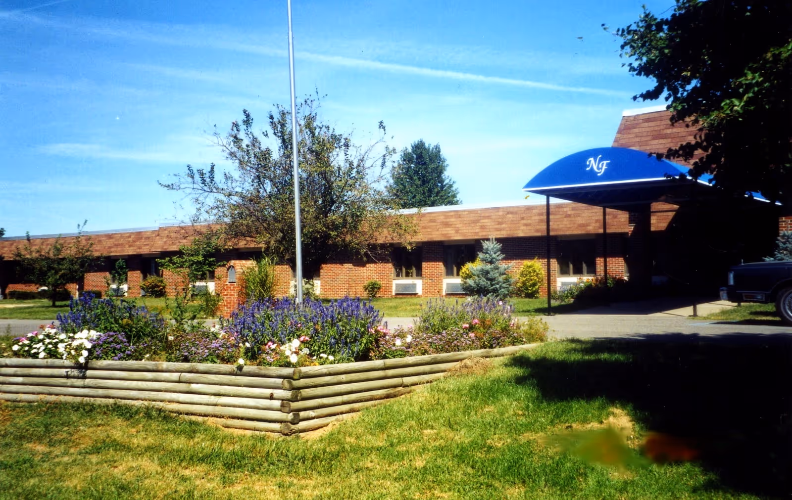 Exterior view of a single-story brick building with a blue awning labeled 'NF' over the entrance. In front of the building is a raised flower bed with various colorful flowers and a small tree. The sky is clear and blue.