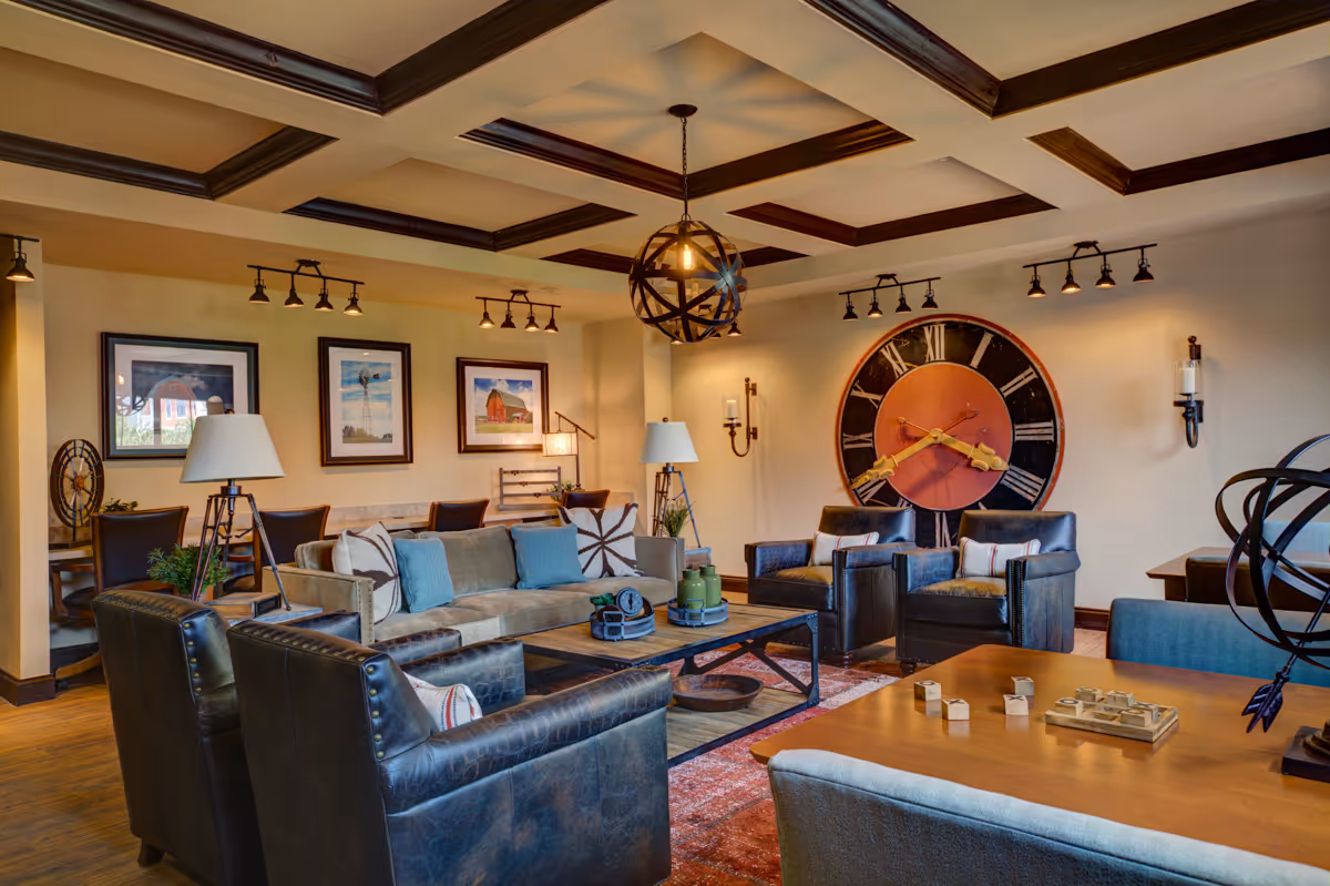 A warm communal living room featuring leather armchairs and sofas around a coffee table, a large decorative wall clock, framed artwork, and a coffered ceiling with pendant lighting.