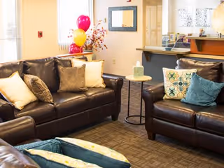 A cozy living room area with dark brown leather sofas adorned with various decorative pillows. There is a small round side table between the sofas with a tissue box on it. In the background, there are colorful balloons and a mirror on the wall, with a reception or service counter visible.