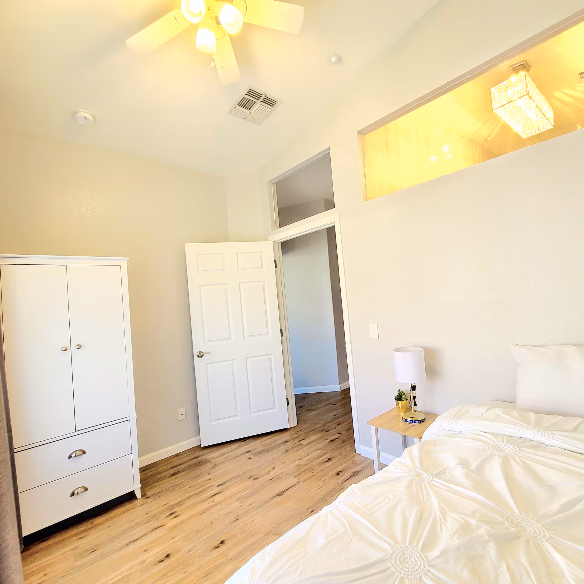 Bright bedroom with a white bed, bedside table and lamp, a white wardrobe, wood flooring, and an open door to a hallway.
