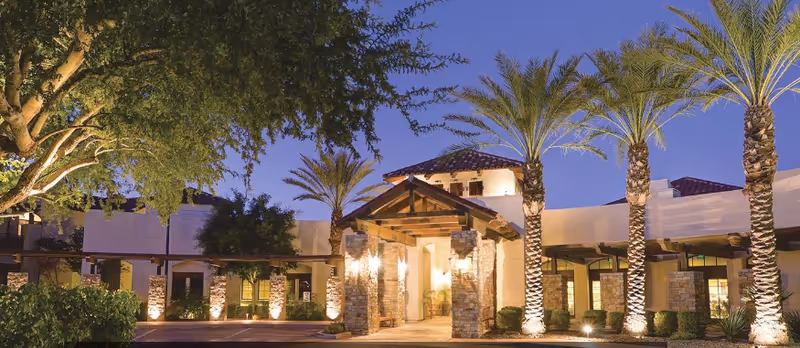 Exterior view of The Village at Ocotillo senior living facility at dusk, featuring a well-lit entrance with stone pillars, palm trees, and landscaped greenery under a clear evening sky.