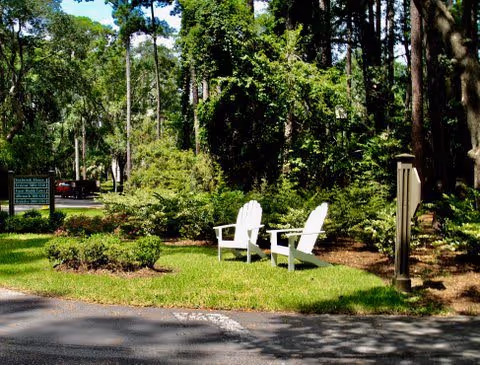 Two white Adirondack chairs placed on a grassy area surrounded by trees and bushes near a paved road, with a sign visible in the background indicating the entrance to The Seabrook of Hilton Head facility.