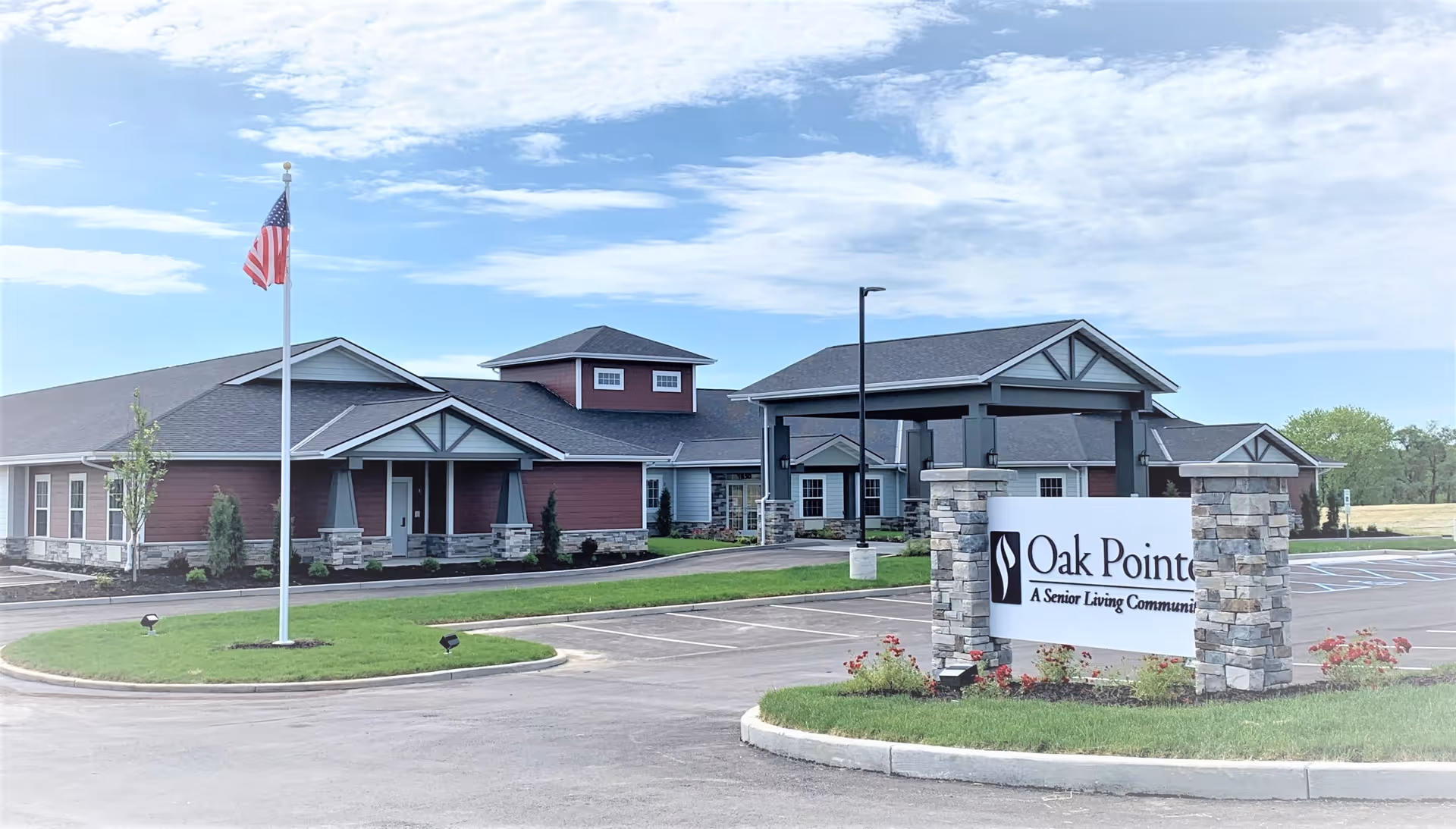 Exterior view of Oak Pointe senior living community building with a parking lot, an American flag on a flagpole, and a sign displaying the facility name. The building has a modern design with stone and wood accents under a partly cloudy sky.