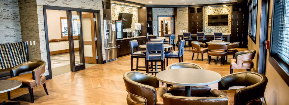 A spacious and well-lit common area in a senior living facility featuring multiple round tables with leather chairs arranged around them. The room has wooden flooring, a kitchenette with a refrigerator and microwave, and a large flat-screen TV mounted on the wall. Large windows with blinds allow natural light to enter the room.